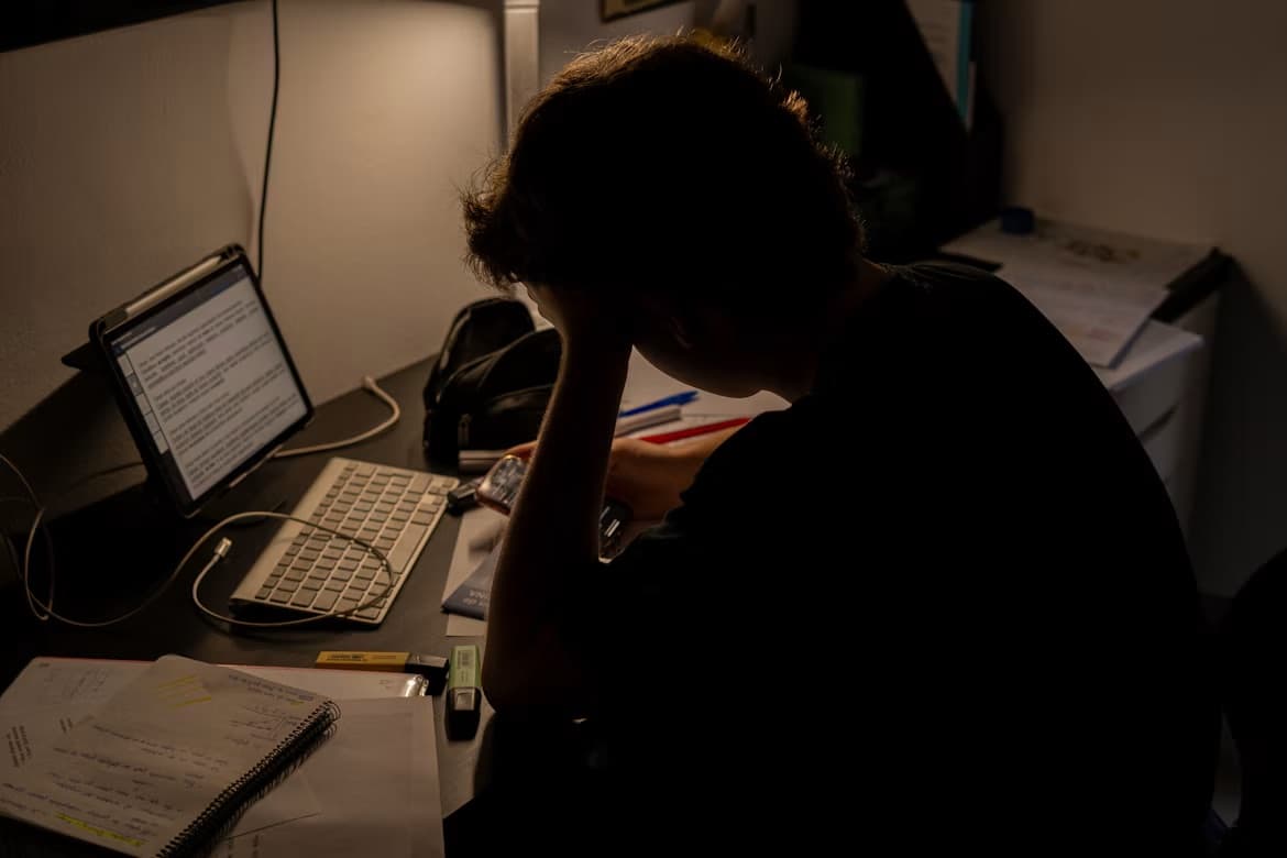 Ung i silhuet sidder ved et skrivebord om aftenen med hånden mod panden, omgivet af noter og en tablet